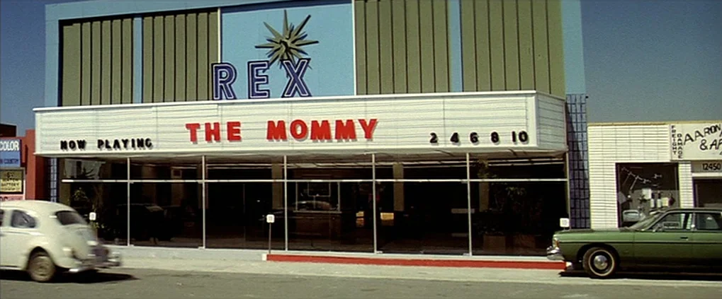 Anguish (1987) movie still: wide — A 1970s Rex cinema building features a marquee displaying the title The Mommy. A vintage white Volkswagen Beetle…