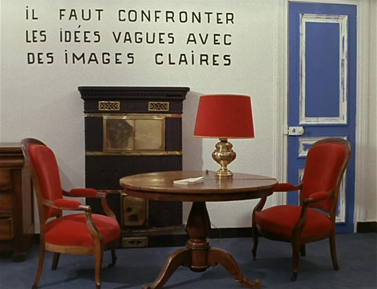 La Chinoise (1967) movie still: wide — A stark room features a white wall with bold black French text painted above. Two matching red armchairs face…