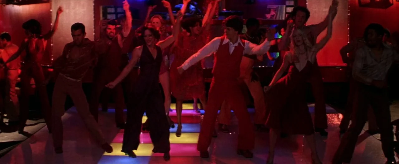 Boogie Nights movie still: wide — A crowded nightclub features a brightly illuminated square grid dance floor with people moving. Many figures dance…