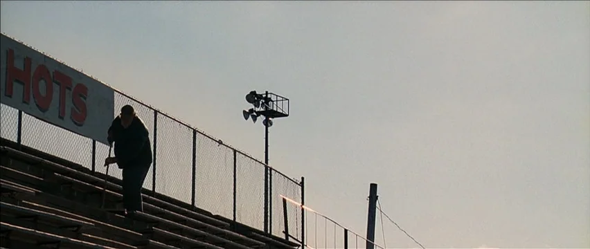 Snow Angels (2007) movie still: wide — A man walks down metal stadium bleachers set against a pale, bright sky. A tall light tower and a chain-link…