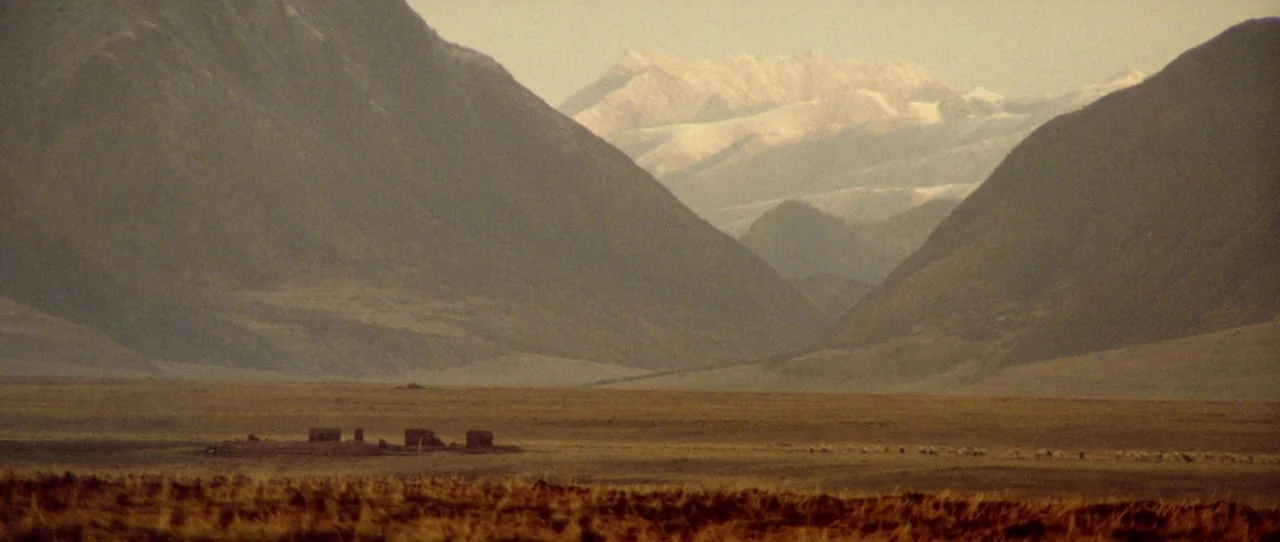 Horse Thief movie still: establishing — A vast, arid high-altitude plateau sits beneath towering, snow-dusted mountains in the distance. A small…