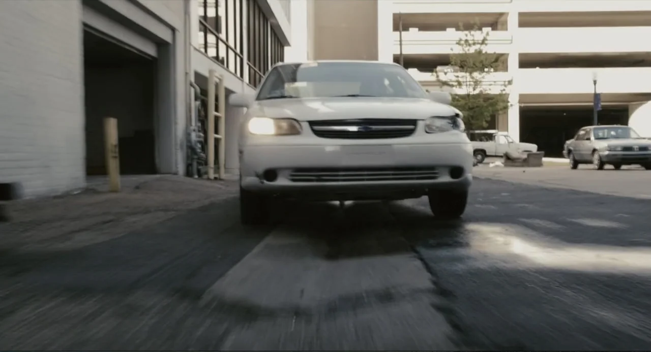 Beyond a Reasonable Doubt (2009) movie still: medium — A white sedan drives toward the lens in a paved parking lot area. Concrete buildings and…