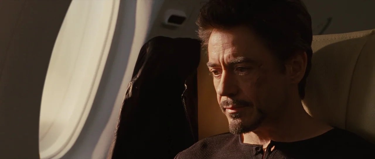 Iron Man 2 (2010) movie still: closeup — A man sits inside the cabin of a private aircraft by a window. He gazes downward with a somber expression…