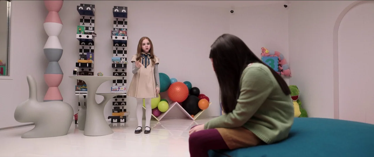M3gan (2022) movie still: wide — A modern, sterile room features white walls, minimalist furniture, and assorted colorful play balls. A doll stands…