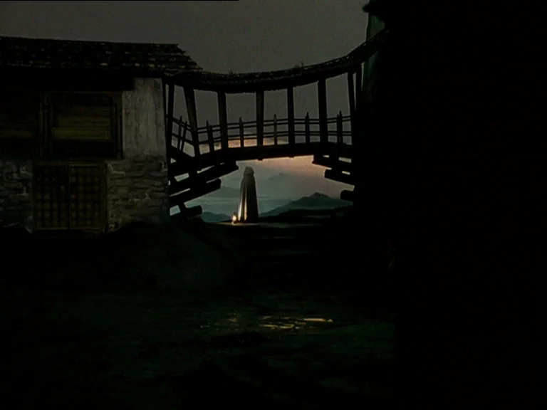 Black Narcissus (1947) movie still: wide — A rustic wooden bridge crosses a gap between dark stone structures at night. A hooded figure stands under…