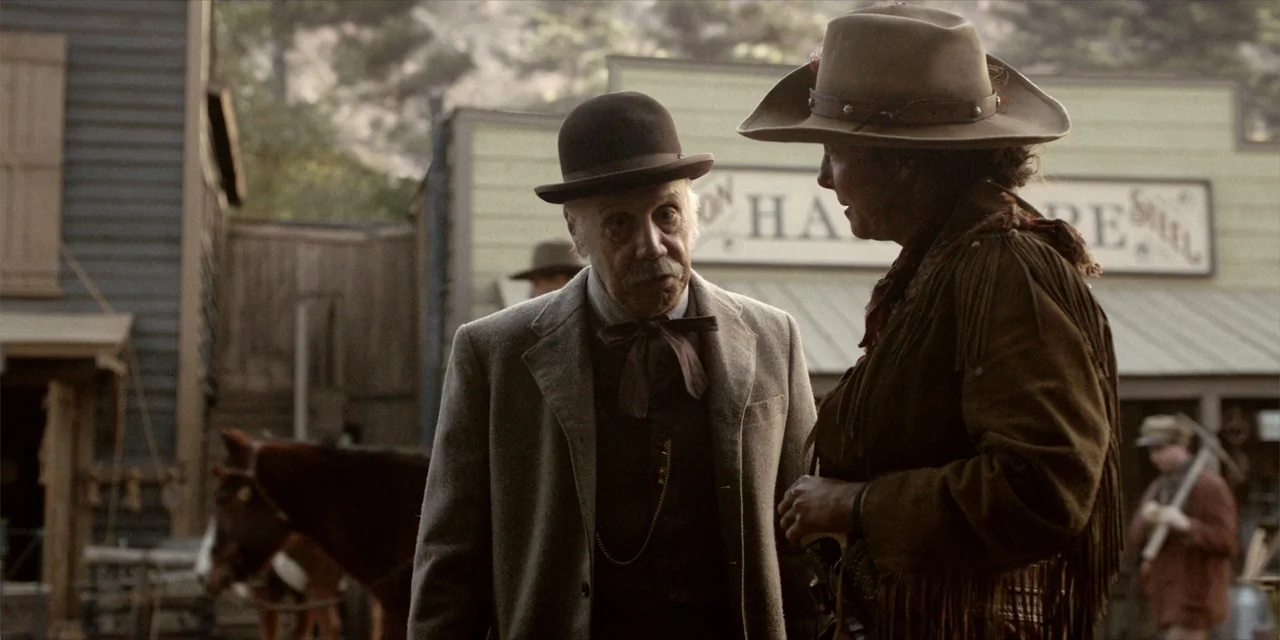 Deadwood: The Movie (2019) movie still: medium — A dusty western town street features wooden buildings and a blurred horse carriage. An older man in…