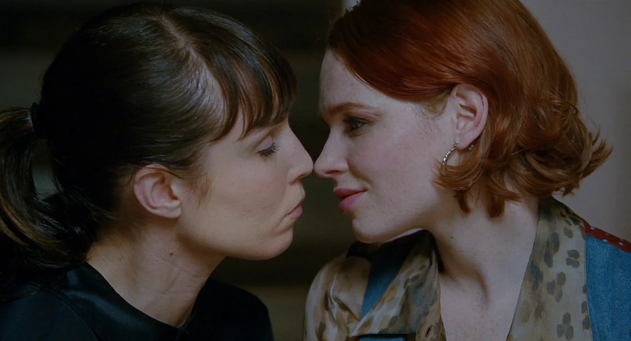 Passion (2012) movie still: closeup — An interior space with indeterminate walls provides a background for two women's faces. The women face each…