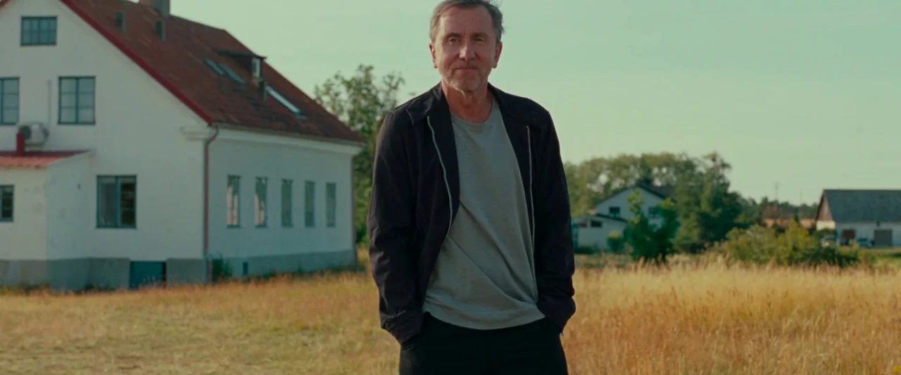 Bergman Island (2021) movie still: medium — An open grassy area sits adjacent to a two-story white house with a red roof. A man stands in the field…