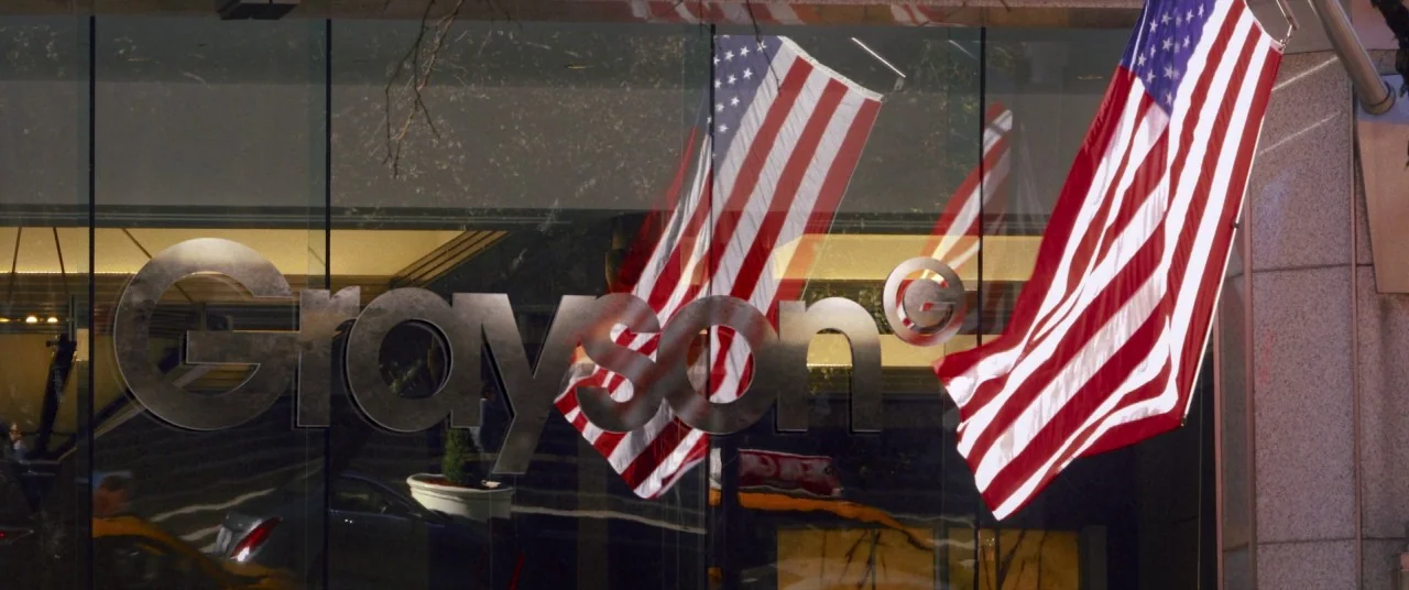 I Care A Lot (2020) movie still: establishing — A glass-fronted office building facade displays the metallic logo for Grayson. American flags are…