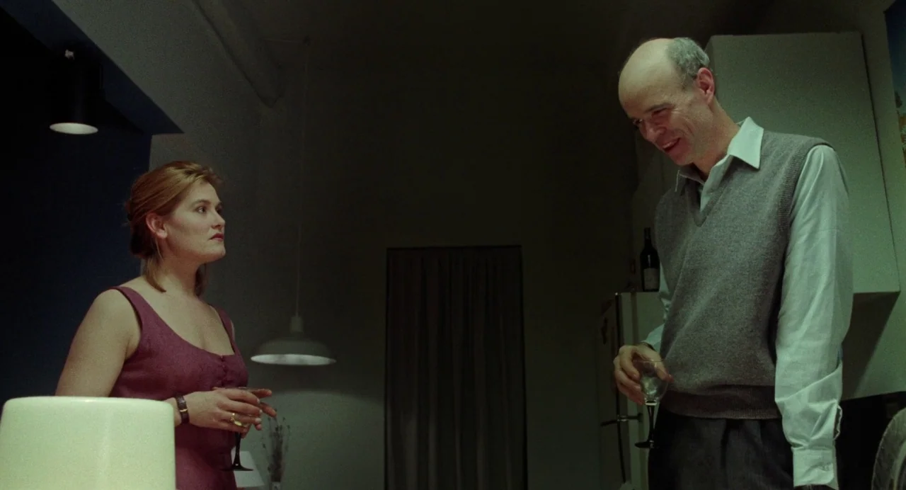 What Happened Was… (1994) movie still: medium — A dimly lit room with muted walls and a dark doorway in the distance. An older man and a younger…