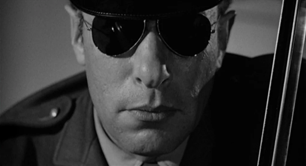 Psycho movie still: closeup — A man in a military cap sits inside a dark vehicle. He wears aviator sunglasses while staring directly ahead.