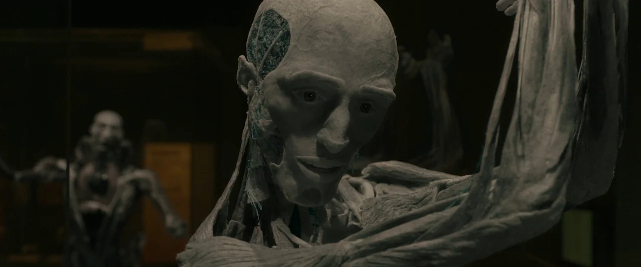 After Yang (2021) movie still: closeup — A dimly lit workshop contains an anatomical android displaying its synthetic musculature. Another similar…