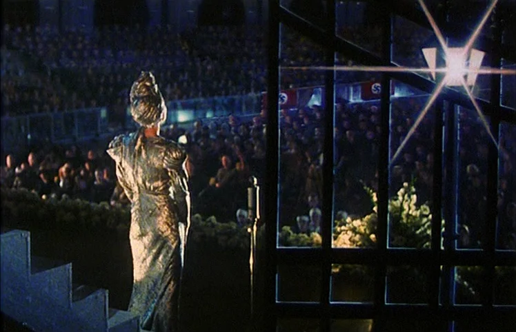 Lili Marleen (1981) movie still: fullbody — A woman in a metallic dress stands on a dark, raised platform overlooking an audience. The crowd sits in…