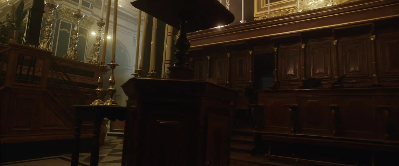 Hounds of War (2024) movie still: establishing — A dark wooden church interior features ornate paneling and carved lecterns under dim light. Haze…