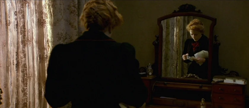 The House of Mirth (2000) movie still: medium — A dimly lit room features a vintage wooden vanity with a large ornate mirror. A young woman with…