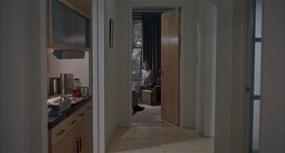 Rosemary’s Baby (1968) movie still: wide — A residential hallway with a kitchen counter to the left and a doorway ahead. A woman sits on a chair in…
