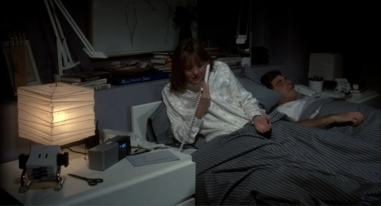 Baby Boom (1987) movie still: medium — A modern bedroom with white furniture and gray striped bedding in a dimly lit room. A woman speaks into a…
