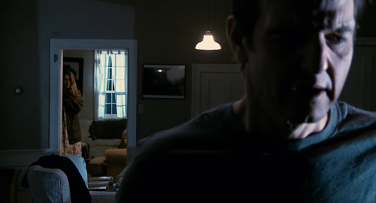 Another Earth (2011) movie still: medium — A dimly lit room is foregrounded by a man in shadow. A woman stands in the frame of an open doorway…