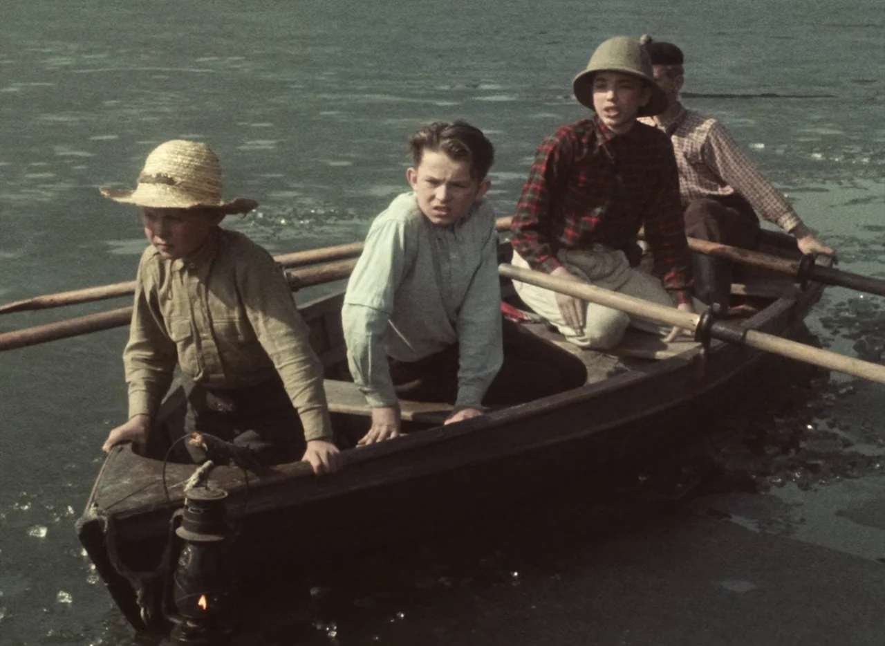 A Journey to the Beginning of Time (1955) movie still: medium — Four boys sit in a small wooden rowboat floating on rippling dark water. The boys…