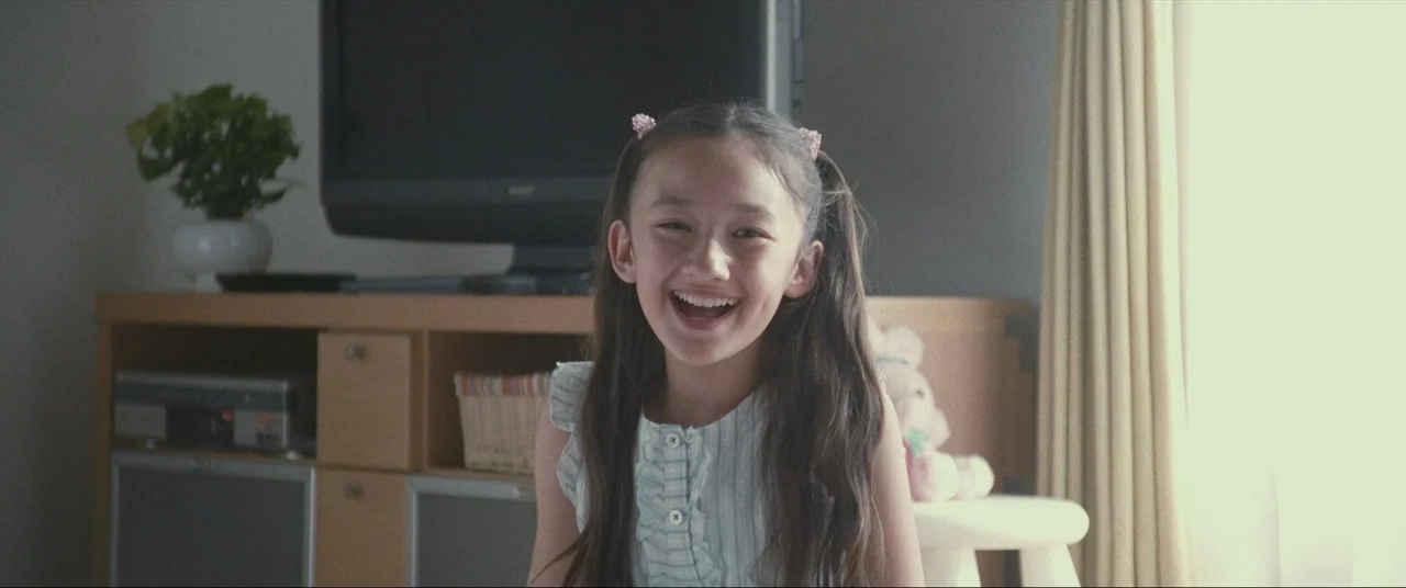 The World of Kanako (2014) movie still: medium — A light-filled room features a television cabinet against a neutral wall. A young girl laughs…