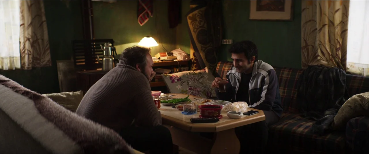 About Dry Grasses (2023) movie still: medium — A dim living room features dark green walls, patterned textiles, and a low central coffee table. Two…