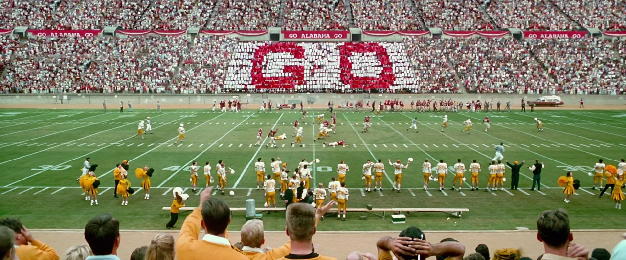 Forrest Gump (1994) movie still: wide — A vast football stadium is packed with spectators holding red and white cards. Players in yellow and white…