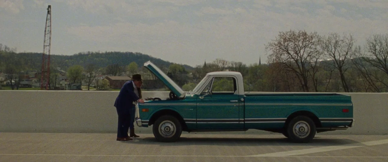 The Old Man & The Gun (2018) movie still: wide — An open-air overpass overlooks a small town nestled in hilly, tree-covered terrain. A man and a…
