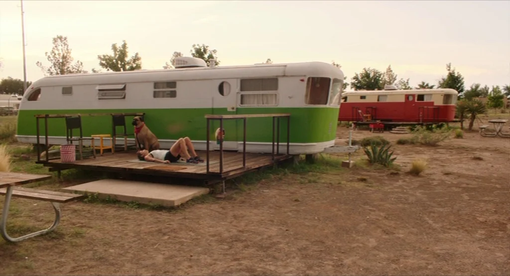 Results (2015) movie still: wide — A rural trailer park features a green-and-white vintage trailer with an attached wooden deck. A woman performs an…