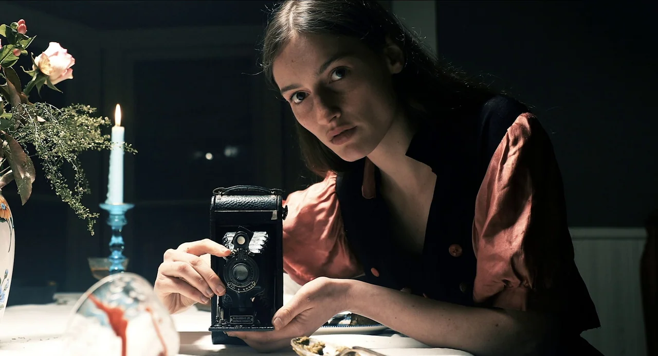 Where The Devil Roams (2023) movie still: medium — A woman sits at a table in a darkened room with a lit candle. She holds a small, black antique…