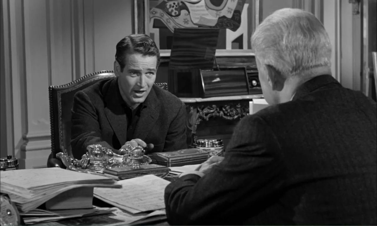 Paris Blues (1961) movie still: medium — A paneled office contains a wooden desk cluttered with papers and ornate metal accessories. Two men in…