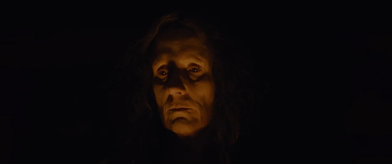 Hagazussa (2017) movie still: closeup — An elderly woman stands in total darkness illuminated only by a low, warm light. Her face is clearly visible…