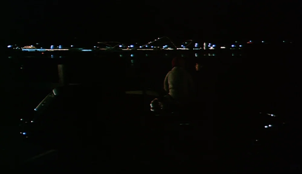 Harold and Maude (1971) movie still: wide — A dark wooden pier extends into a black body of water at night. A man sits on the edge, gazing at…