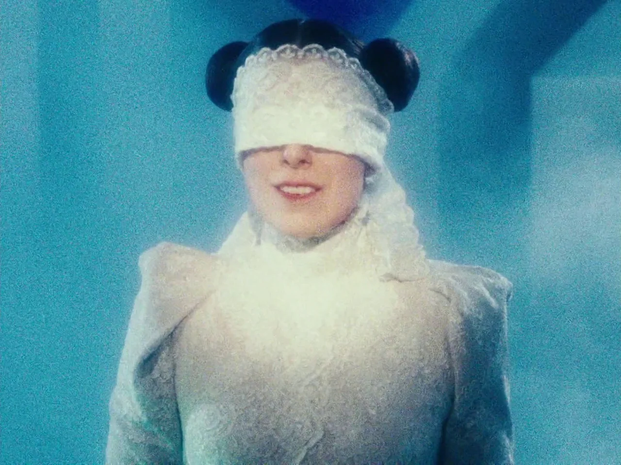 The Twentieth Century (2019) movie still: medium — A woman wears a white lace blindfold against a solid light blue background. She stands facing the…
