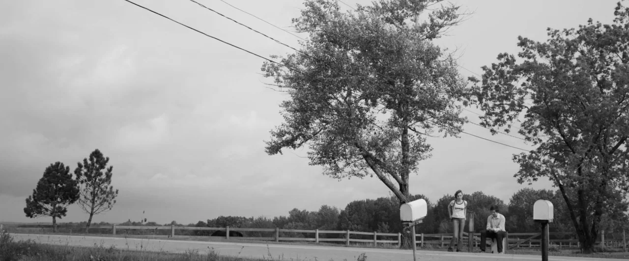 Weirdos (2016) movie still: wide — A rural roadside with a wooden fence, two mailboxes, and tall leafy trees. A young woman stands while a young man…