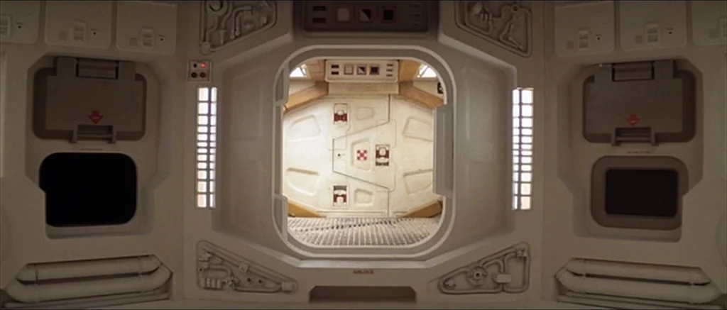 Alien (1979) movie still: establishing — A stark white modular spaceship airlock features a central doorway and heavy mechanical bulkheads. The…