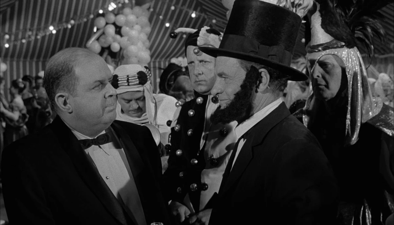 The Manchurian Candidate (1962) movie still: medium — A crowded tent filled with people in various elaborate costumes at a party. A balding man in a…