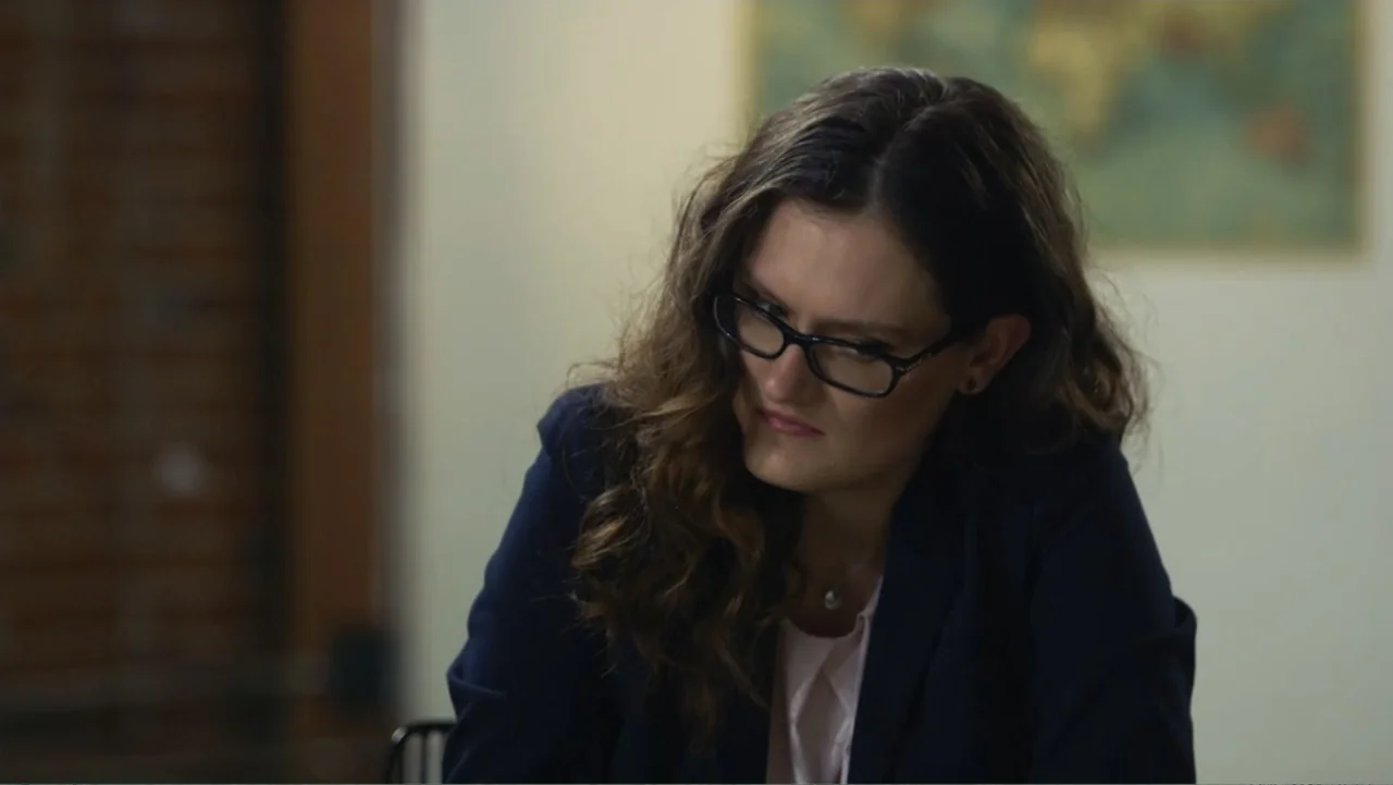 Finding Amelia (2024) movie still: medium — An office interior features a blurred map on a wall and a brick pillar. A woman with long wavy brown…