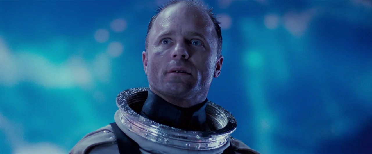 The Abyss (1989) movie still: closeup — A man in a spacesuit looks upward against a swirling blue background. The metallic collar of his suit…
