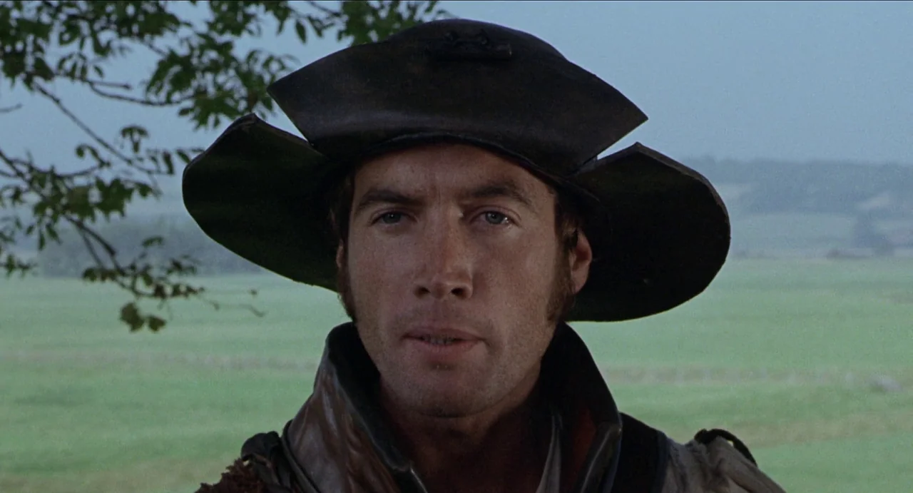 The Canterbury Tales (1972) movie still: closeup — An open green field stretches out behind a man under an overcast sky. A man in a leather hat and…
