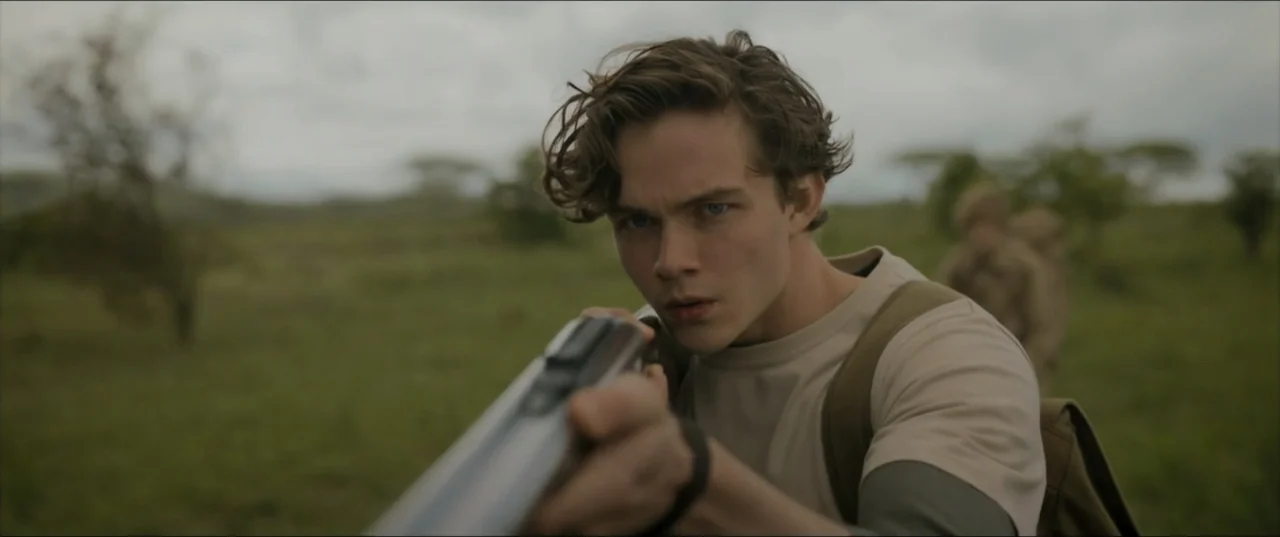 Kraven the Hunter (2024) movie still: medium — A grassy savanna with scattered bushes under an overcast sky. A young man holds a shotgun forward…