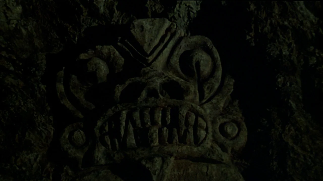 Sorcerer (1977) movie still: closeup — A stone relief carving is set into the dark, rough surface of a cave. The carving displays an anthropomorphic…
