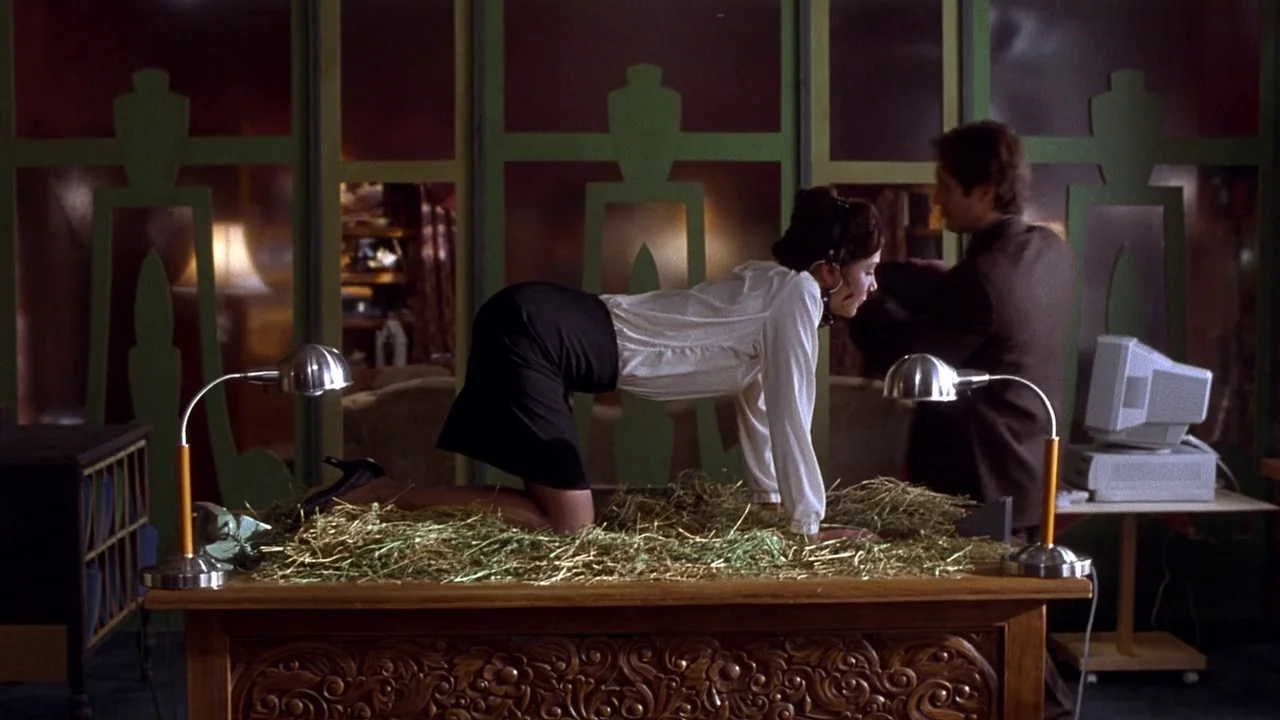 Secretary movie still: medium — An office environment features green paneled walls with abstract patterns and a wood desk. A woman moves across the…