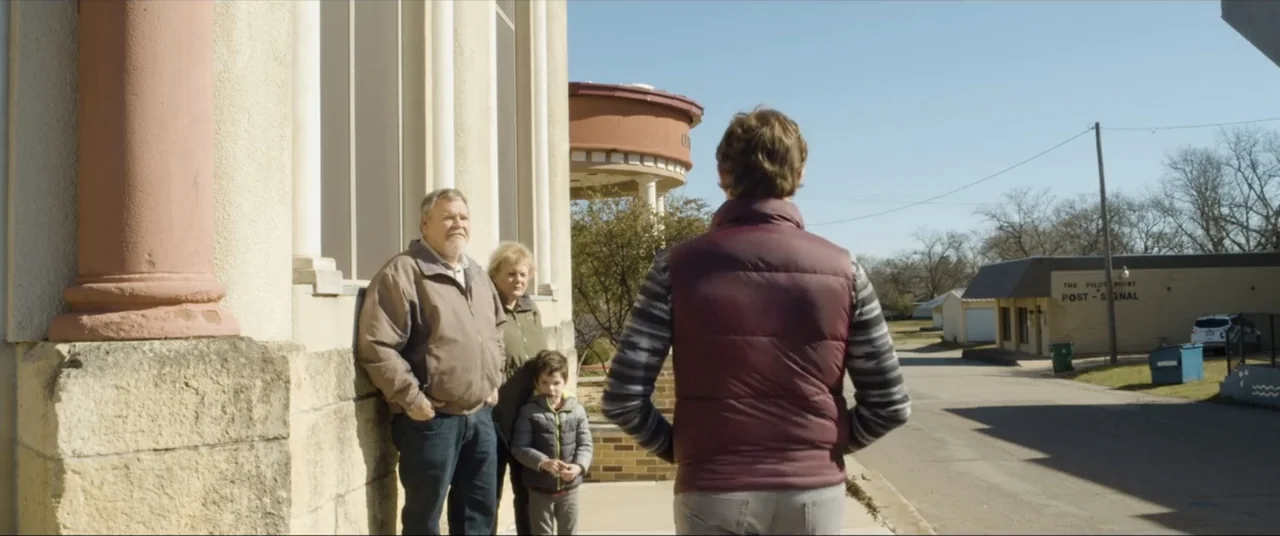 Amityville Cult (2021) movie still: fullbody — A group of people stands on a sidewalk next to a stone building exterior. One man faces an older…