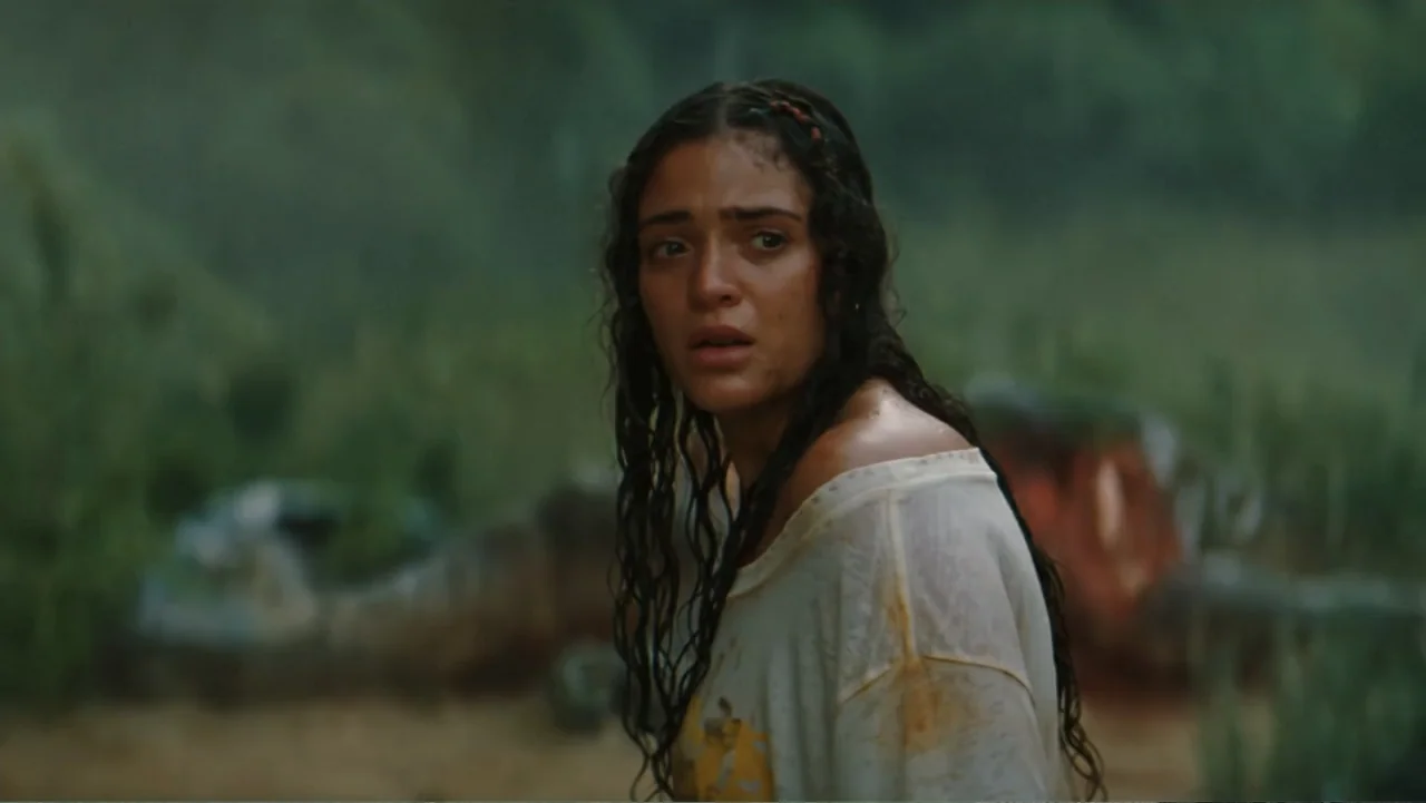 Jurassic World Rebirth (2025) movie still: medium — A dense green jungle setting with blurred foliage and patches of brown earth. A young woman with…
