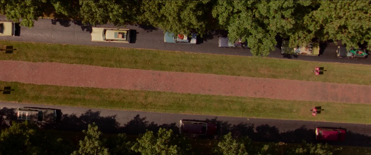 White Noise (2022) movie still: establishing — An overhead view shows a long straight road divided by a red brick median. Various passenger cars…