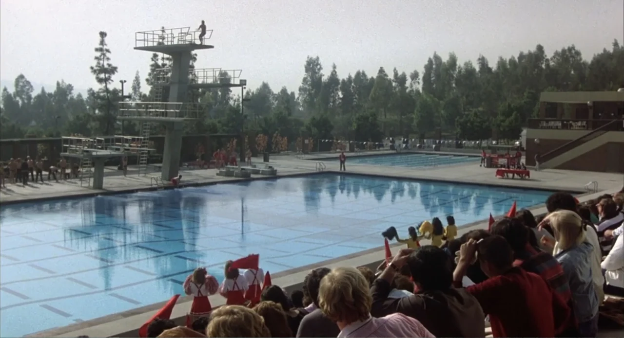 Back to School (1986) movie still: wide — An outdoor aquatic facility features a large competition pool and a tall diving platform. A crowd of…