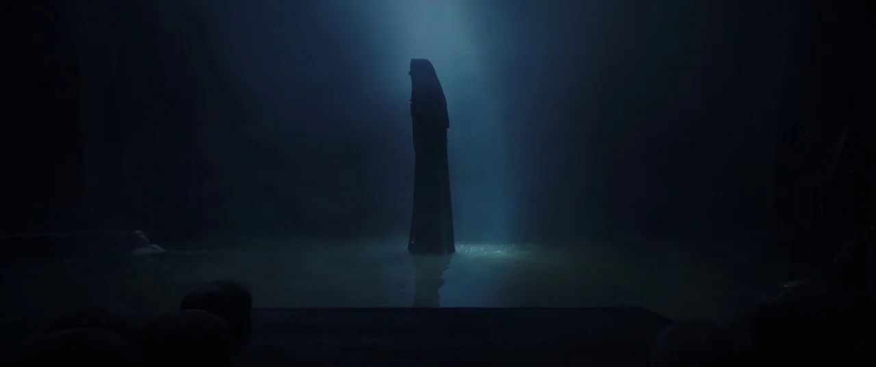 The Nun (2018) movie still: wide — A dark, cavernous space contains a pool of shallow, still water. A hooded figure stands perfectly still in the…