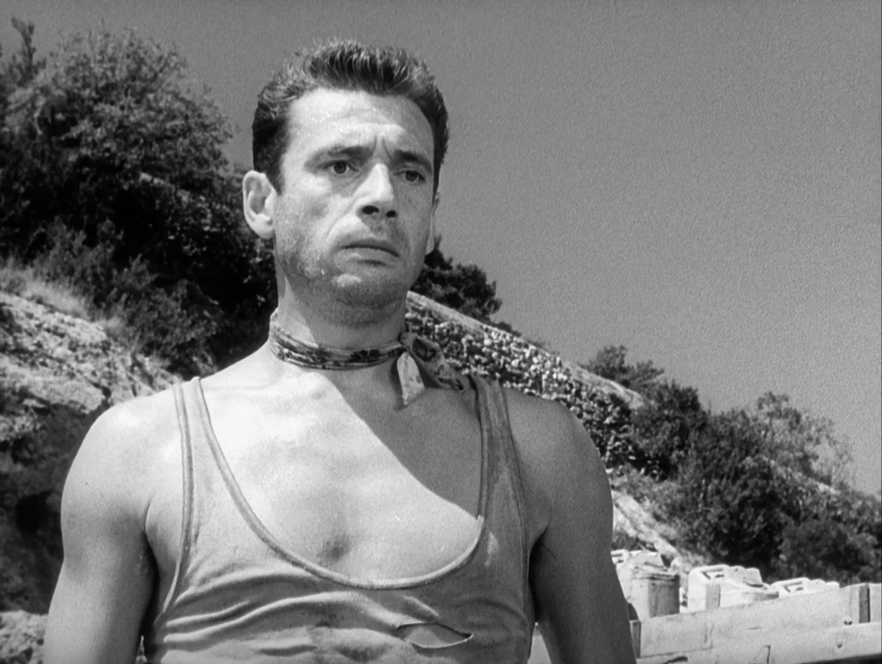 The Wages of Fear (1953) movie still: medium — A rocky, barren coastal environment with sparse vegetation under a clear, bright sky. A man stands in…