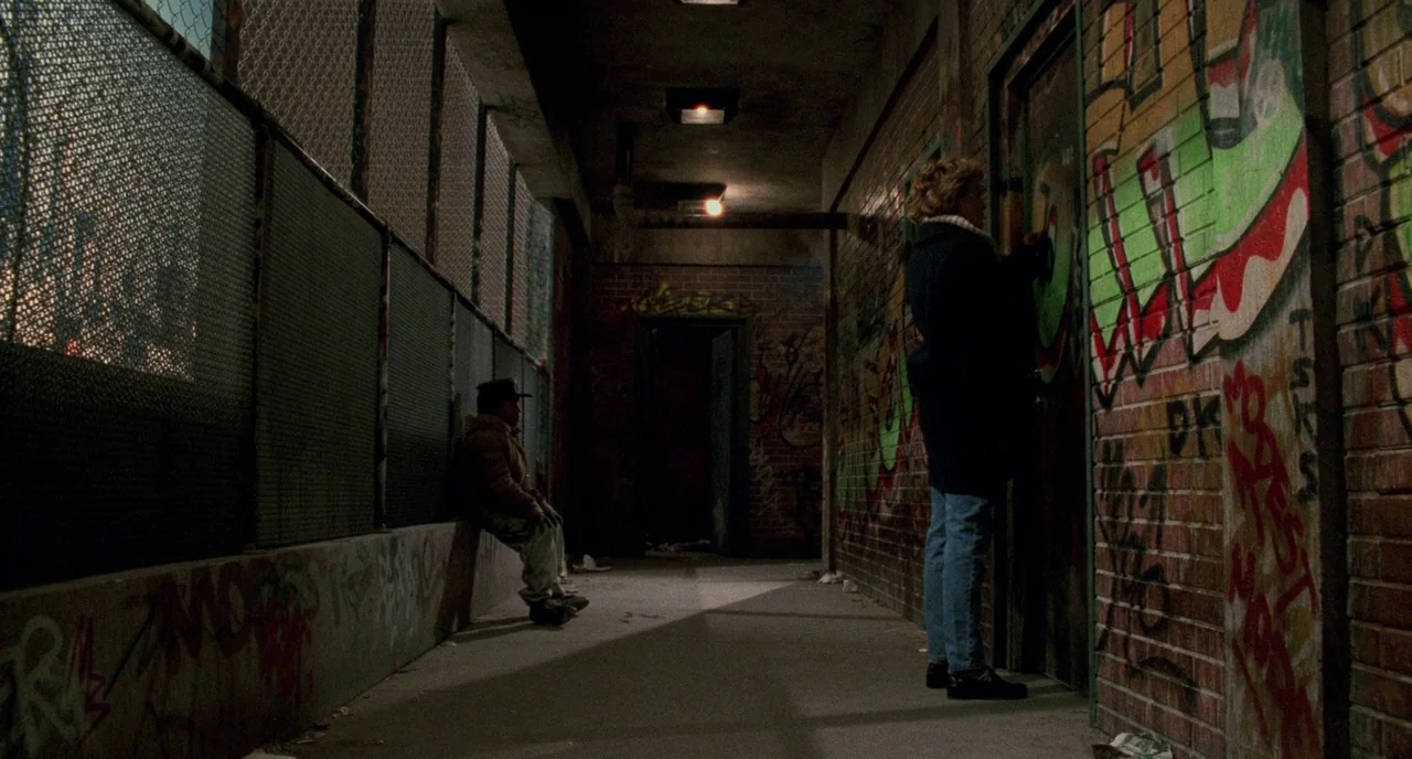 Candyman (1992) movie still: wide — A narrow exterior concrete walkway is enclosed by high chain-link fencing and graffiti-covered brick walls. One…