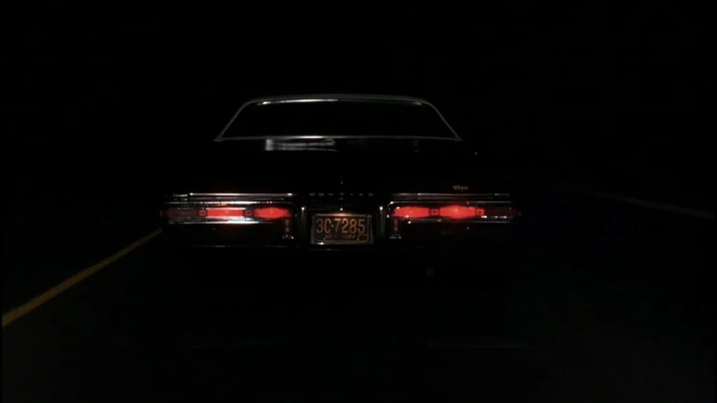 Goodfellas (1990) movie still: medium — The rear of a black vintage sedan moves along a dark, asphalt road at night. Bright red rectangular…